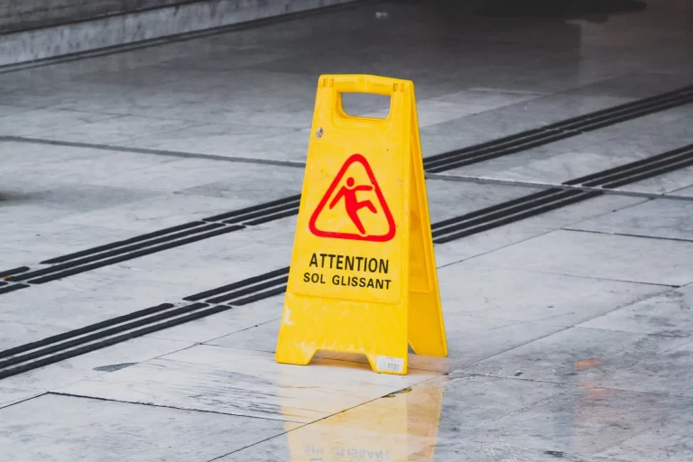 Panneau jaune signalant un sol glissant, posé sur un revêtement mêlant blanc, gris et noir.