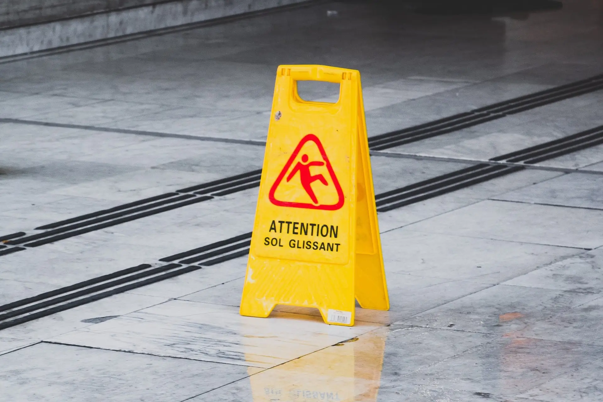 Panneau jaune signalant un sol glissant, posé sur un revêtement mêlant blanc, gris et noir.