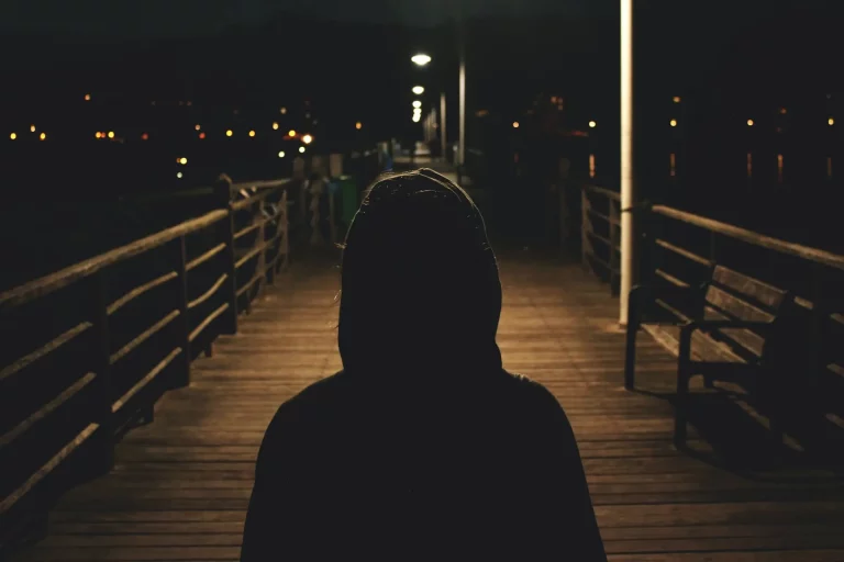 Une silhouette, capuche relevée, se tient sur un pont plongé dans l'obscurité de la nuit.