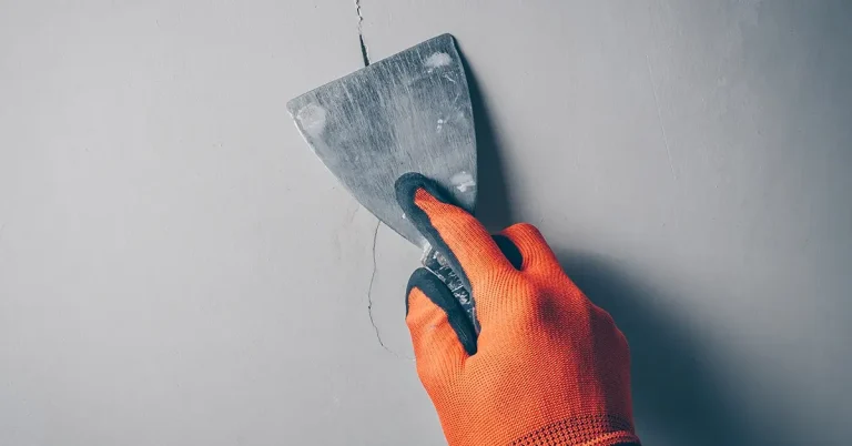 Une main gantée en orange tient une spatule pour réparer une fissure dans un mur.
